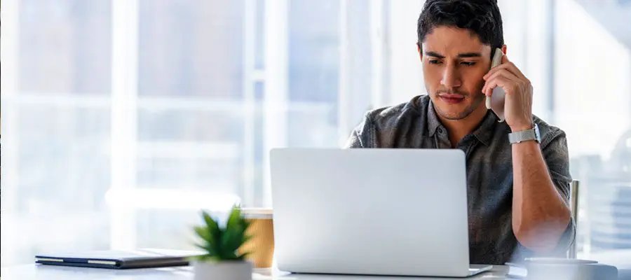 Focused man on a phone call troubleshooting an issue on a laptop at a bright desk.